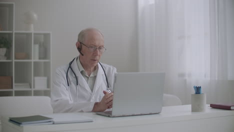 Ein-älterer-Männlicher-Arzt-Berät-Patienten-Online,-Sitzt-Im-Büro-Der-Klinik-Und-Kommuniziert-über-Das-Internet-Auf-Einem-Laptop